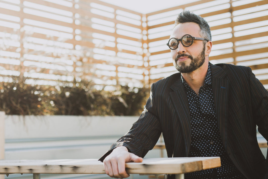 man on patio wearing suit jacket and round black wood glasses on face. 